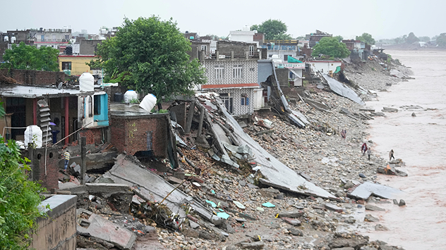 Houses on the riverbank have been severely damaged by flooding and landslides, debris is scattered everywhere, and people are walking along the river.